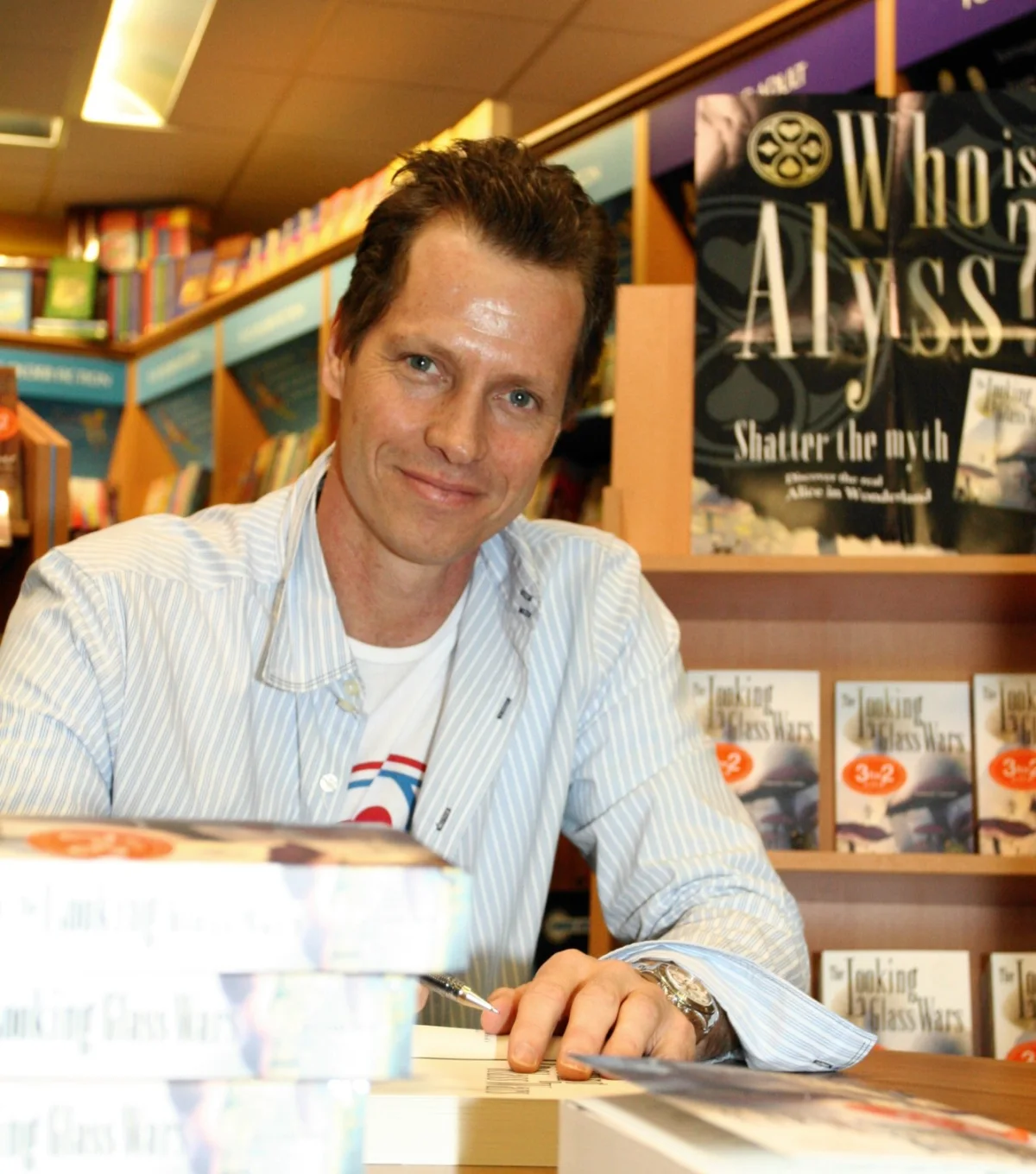 Frank Beddor at a signing table in a bookshop, looking up at
                  the camera with a pen in his hand and an open copy under it,
                  a stack of The Looking Glass Wars at his elbow. Behind him a
                  tall display reads &ldquo;Who is Alyss? Shatter the myth
                  &mdash; discover the real Alice in Wonderland&rdquo;, over
                  shelves of the paperback under 3 for 2 stickers.