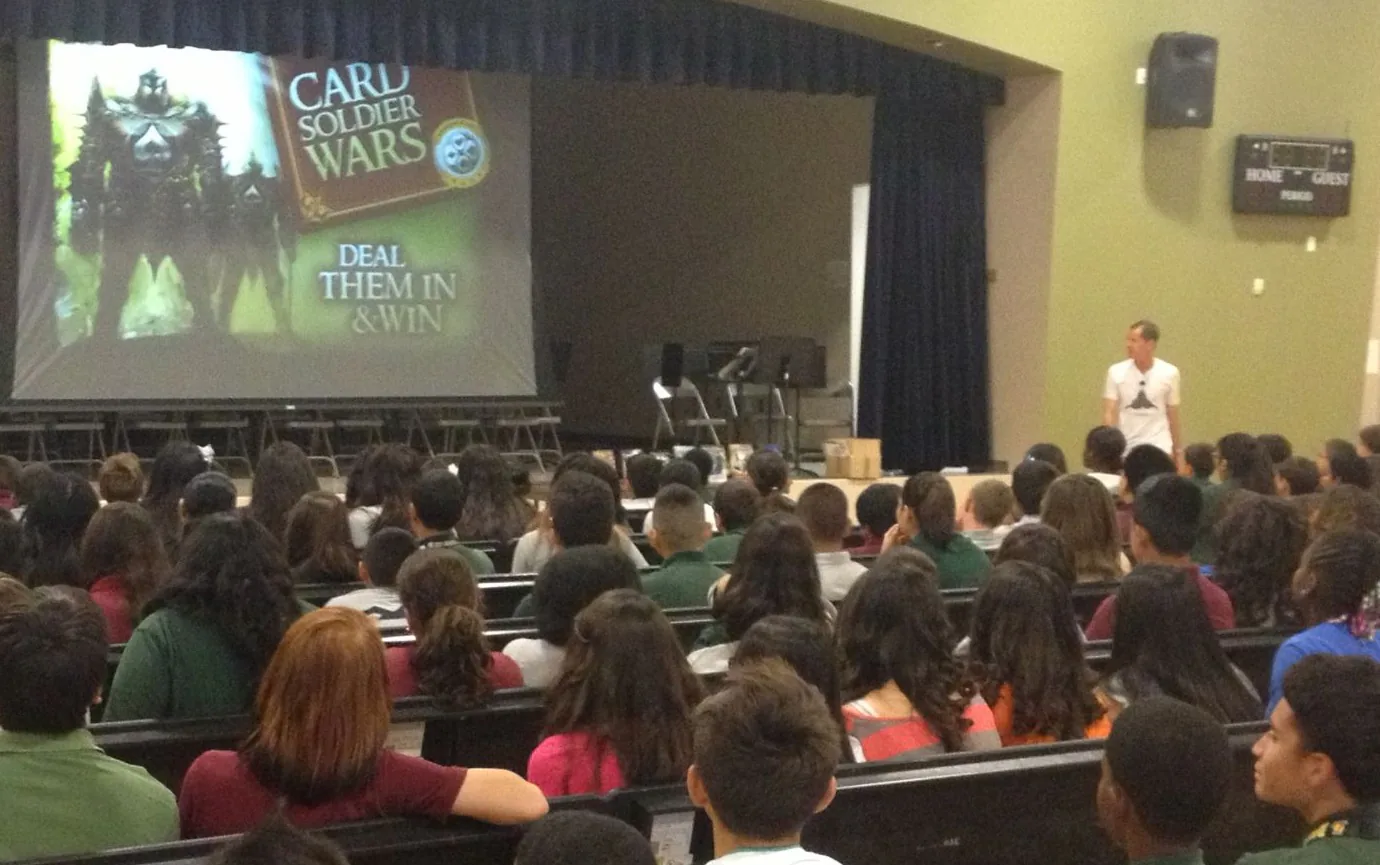 Beddor at the front of a packed school auditorium, a screen
               behind him showing Card Soldier Wars &mdash; Deal Them In &
               Win, rows of students watching from the benches.