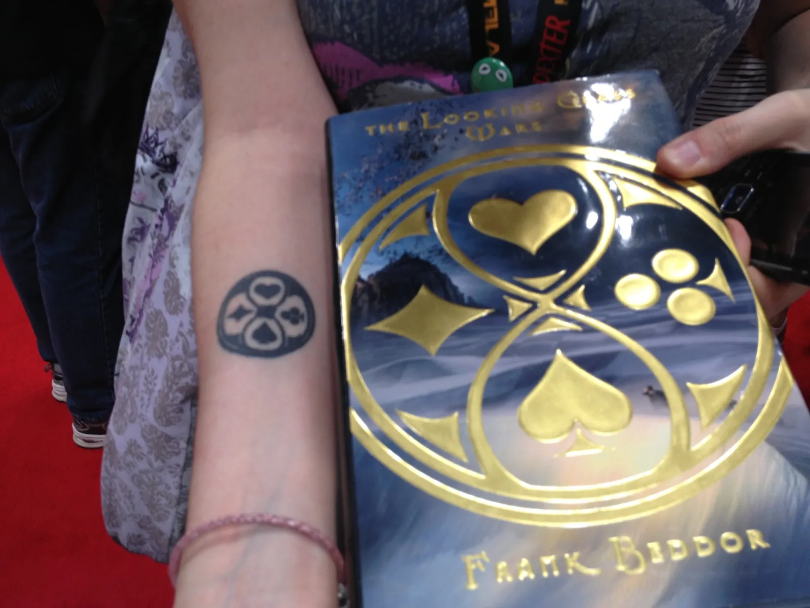 A fan&rsquo;s forearm tattooed with the four-suit emblem, held
               beside a copy of The Looking Glass Wars whose gold-foil cover
               carries the same emblem.