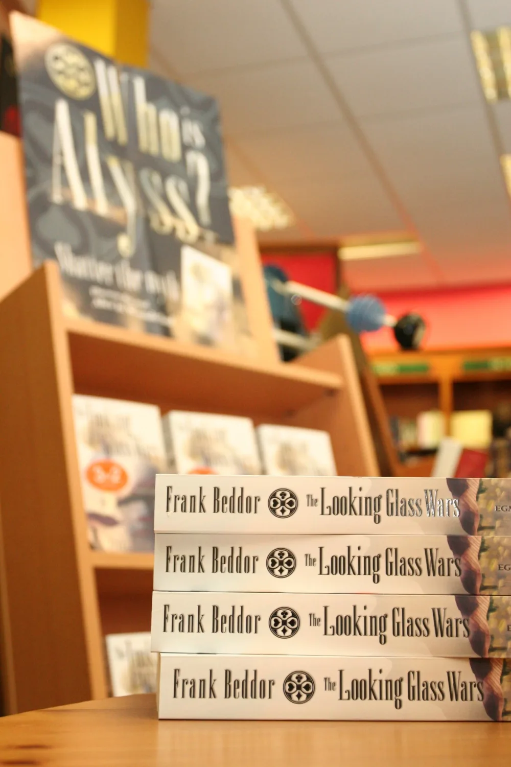 Four copies of The Looking Glass Wars stacked spine-out on a
                    bookshop table, Frank Beddor&rsquo;s name and the Wonderland
                    card-suit emblem running along each one. Behind them, out of
                    focus, the Who is Alyss? display and a shelf of the same
                    paperback under 3 for 2 stickers.