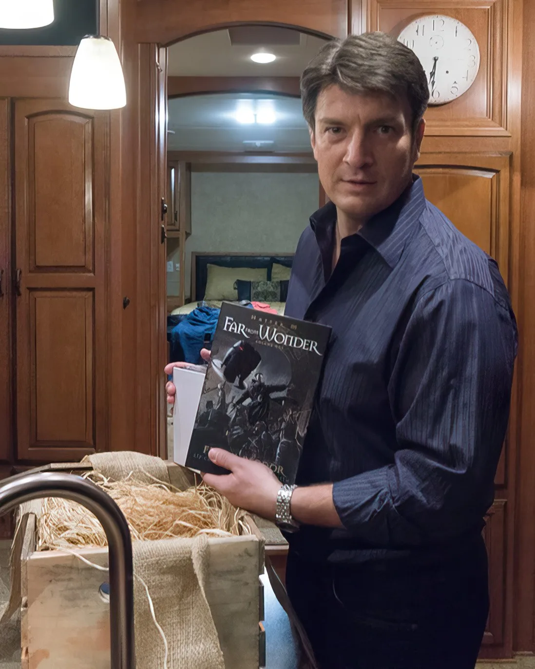 Nathan Fillion in a navy shirt holding up a copy of Hatter M:
               Far from Wonder beside a straw-packed wooden crate, a wall clock
               and wood-panelled cabinets behind him.