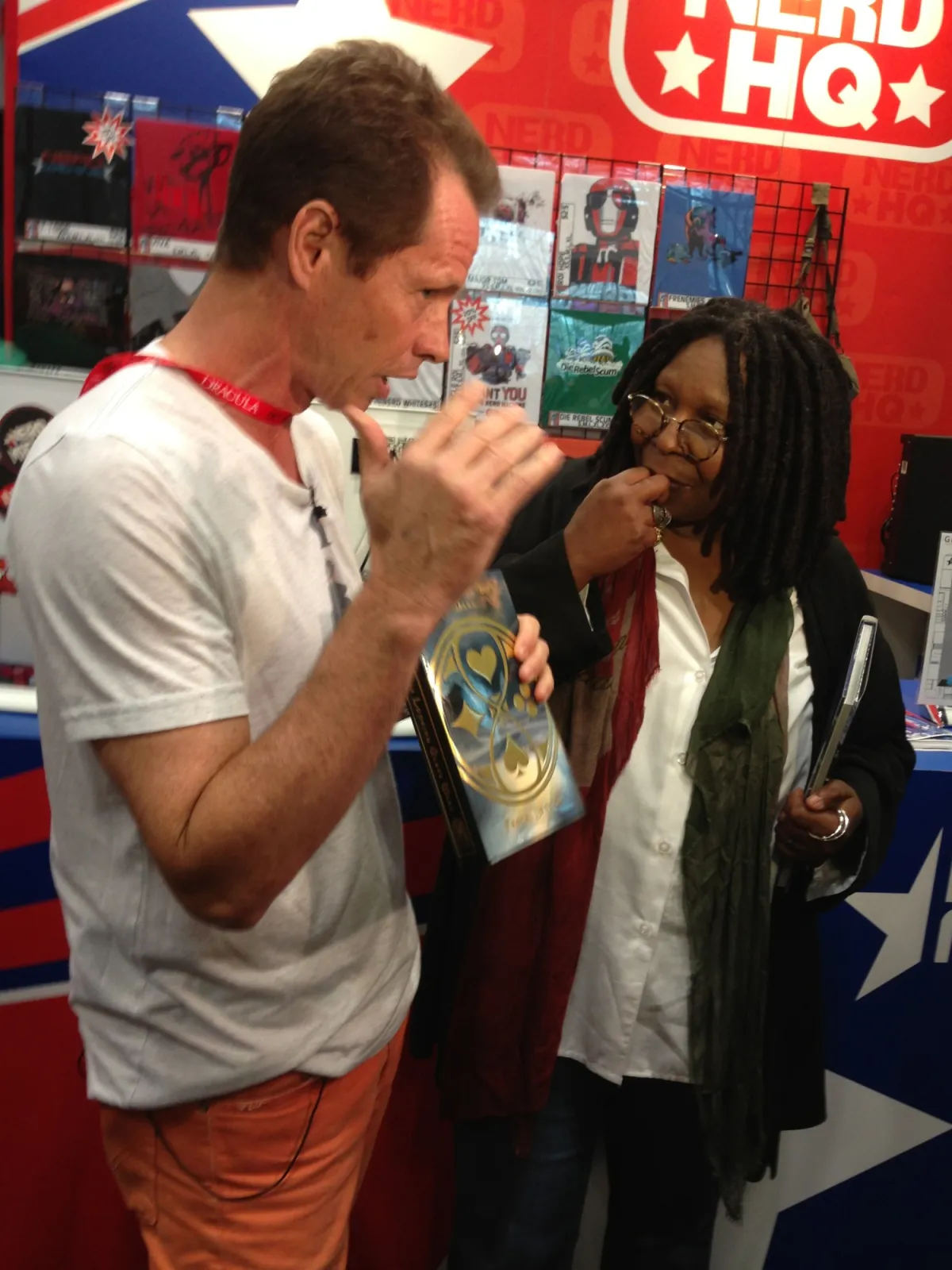Beddor in a white T-shirt and orange trousers at the Nerd HQ
               booth, talking with his hands, holding a copy of The Looking
               Glass Wars, while a woman in round glasses and dreadlocks
               listens with a hand to her chin, a notebook under her arm.