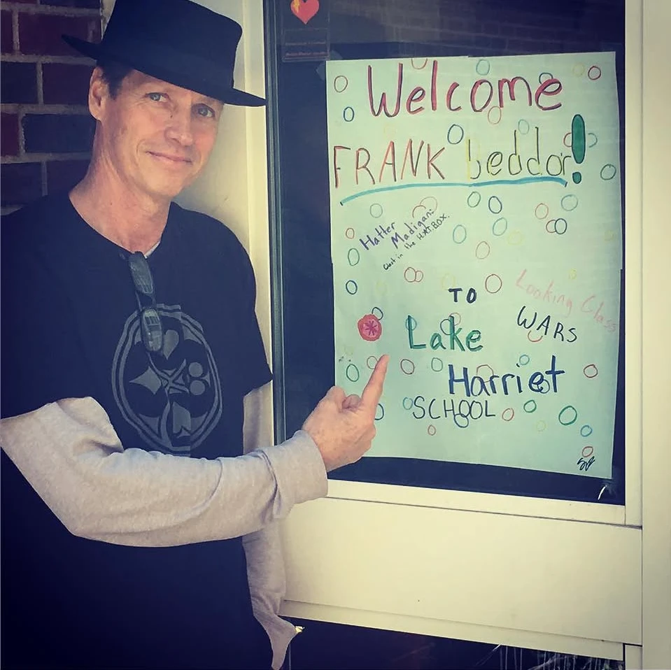 Beddor in a black hat and a Looking Glass Wars T-shirt
               pointing at a hand-drawn poster in a school door: Welcome Frank
               Beddor to Lake Harriet School, with the titles of his books
               written around it.