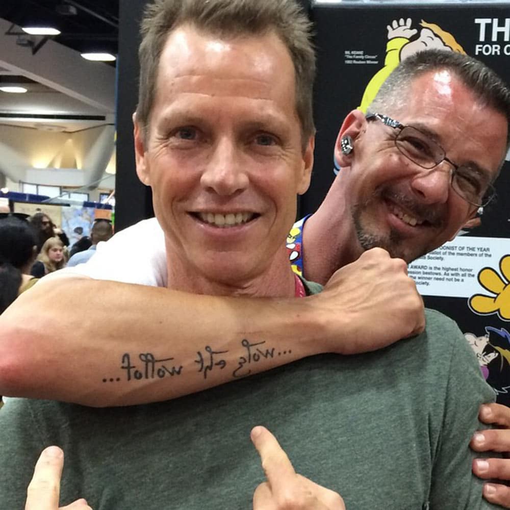 A grinning fan with his arm round Beddor&rsquo;s shoulders at
               Comic-Con, his forearm tattooed &hellip;follow the glow&hellip;
               with the last two words written mirror-image, as Beddor
               points up at it.
