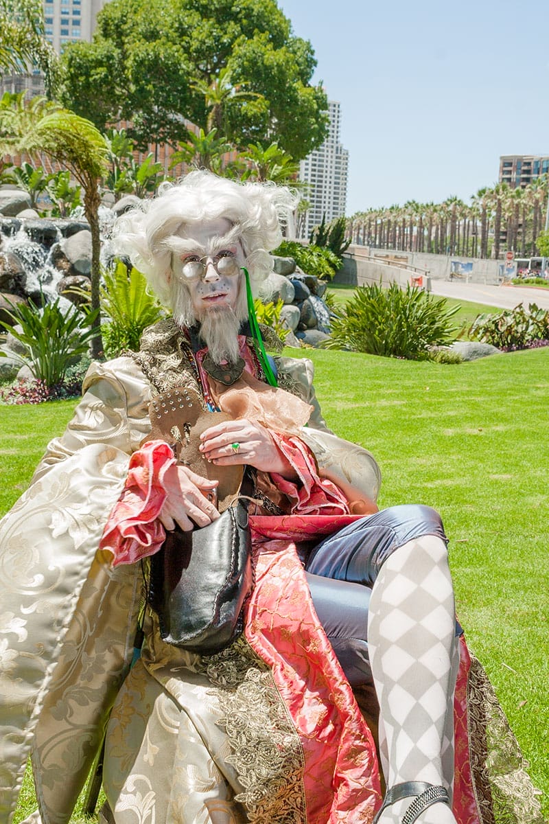 A Bibwit Harte cosplayer seated on a lawn under the San Diego
               sun, in white wig, round spectacles and a cream
               brocade robe lined in red, harlequin stockings
               crossed.