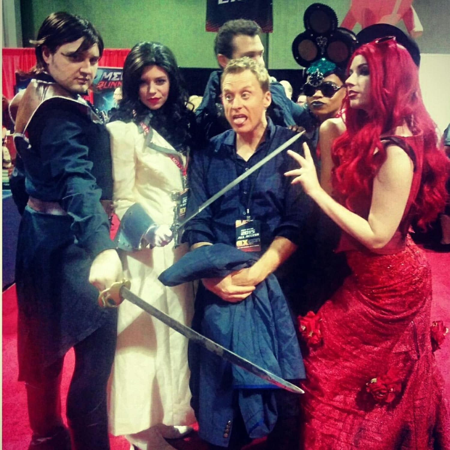 Costumed Looking Glass Wars characters crowding round a grinning
               man at a convention: a swordsman in a dark coat,
               a dark-haired woman in white, and a red-haired
               Redd in a crimson rose gown.