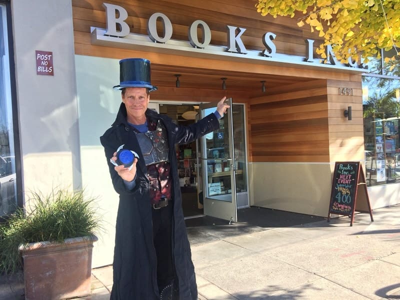 Beddor on the pavement outside Books Inc in Hatter
               Madigan&rsquo;s costume &mdash; long black coat, breastplate and blue
               top hat &mdash; holding out a prototype of the folded hat in one hand
               and pointing at the shop door with the other.