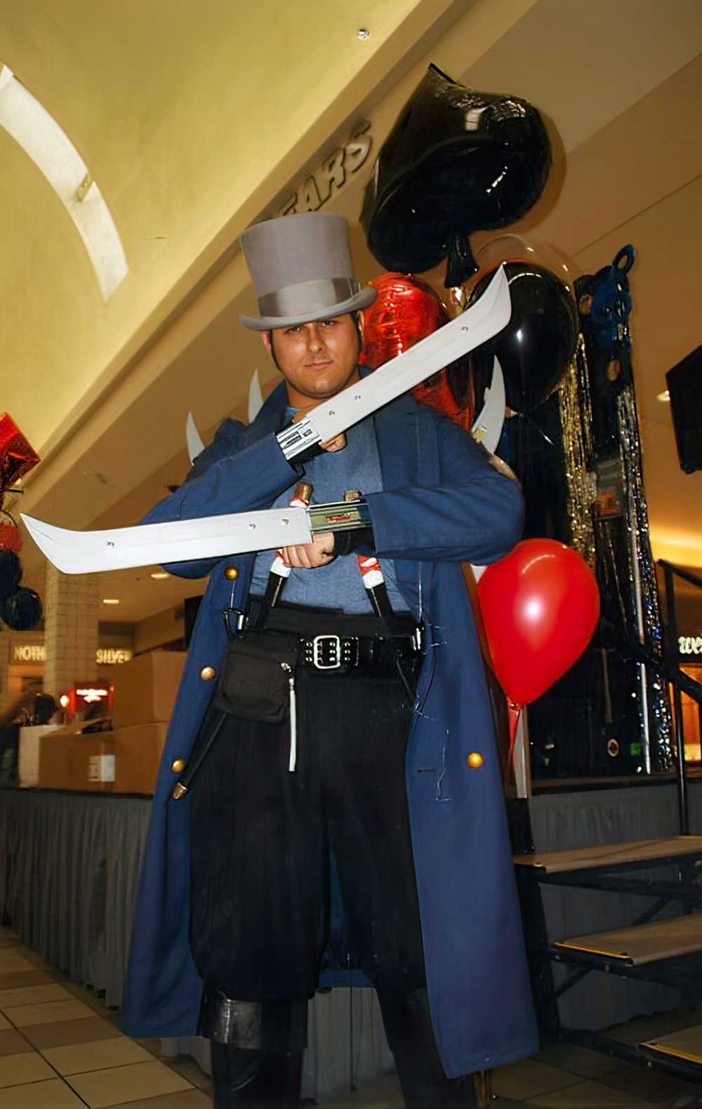 A Hatter Madigan cosplayer in a grey top hat and long blue coat,
               a curved blade in each hand, in a shopping-centre
               atrium hung with black spade and red heart
               balloons.