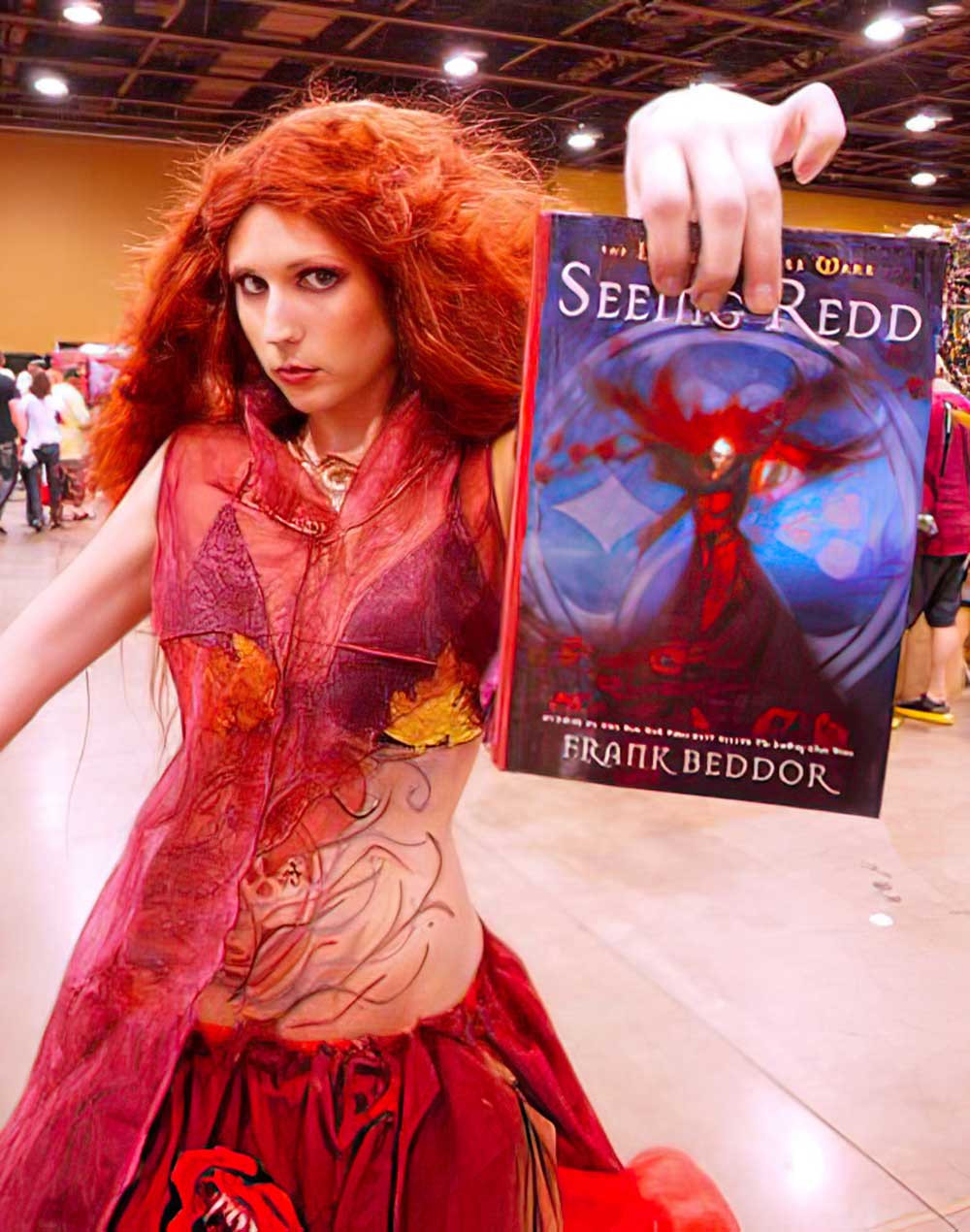 A Redd cosplayer with a wild mane of red hair and a painted bare
               midriff, holding a copy of Seeing Redd out at the
               camera on a convention floor.