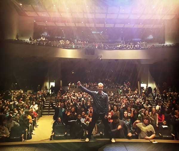 Beddor on the edge of a stage with one fist raised, a full
               auditorium of students behind him waving and cheering.
