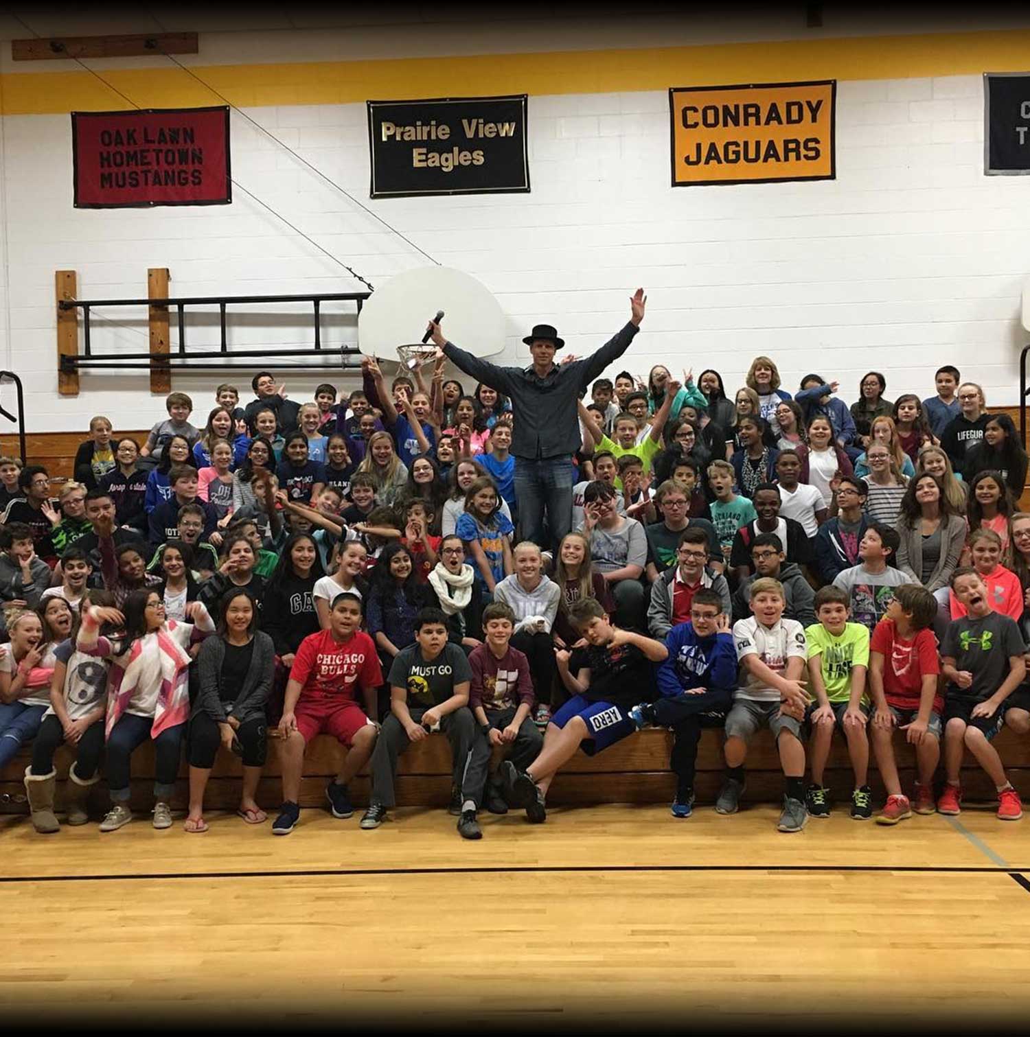 Beddor in a black hat, arms flung wide, standing among a
               packed school gym of cheering students on the bleachers.