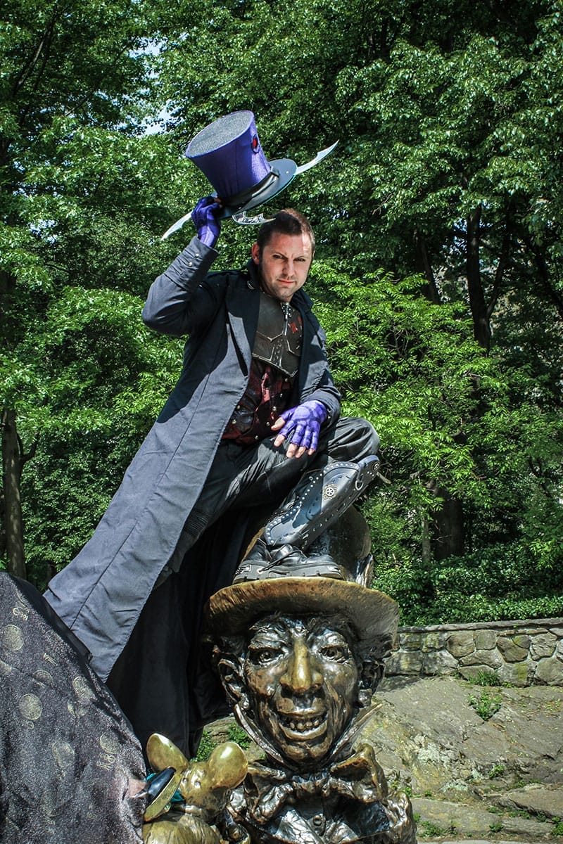 A Hatter Madigan cosplayer in a long grey coat and purple
               gloves, perched on the bronze Mad Hatter of the
               Alice statue in Central Park and raising his
               blade-rimmed top hat.