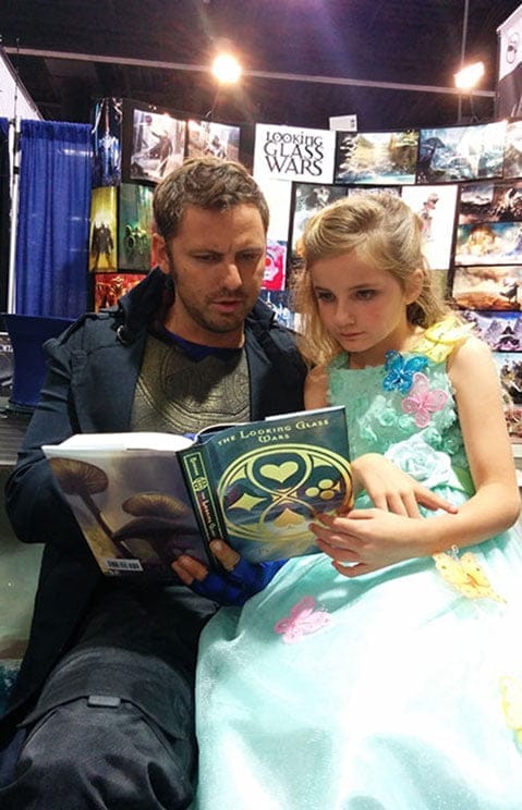 A Hatter Madigan cosplayer at the Looking Glass Wars booth
               reading the book with a young girl in a pale green party dress
               sitting beside him.