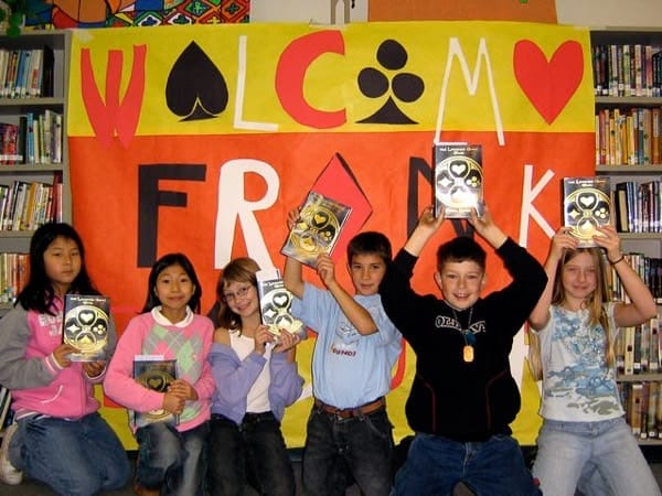 Six schoolchildren in a library holding up copies of The Looking
               Glass Wars in front of a hand-painted banner
               reading Welcome Frank, lettered with card suits.