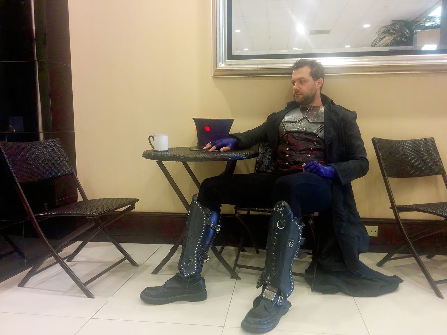 A Hatter Madigan cosplayer in armoured breastplate, long coat
               and studded boots, resting at a cafe table with a mug, his
               red-badged top hat on the table beside him.