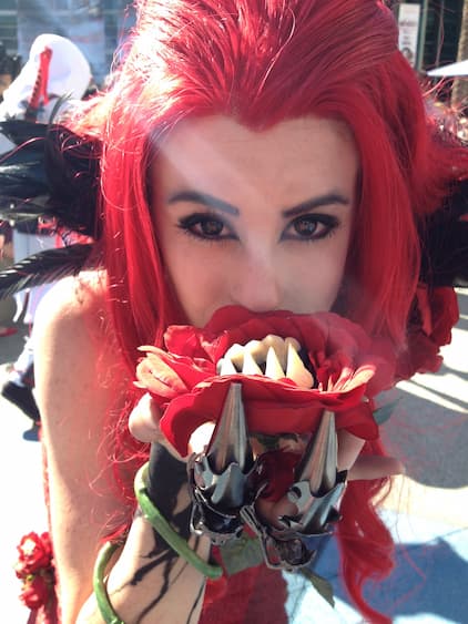 A Redd cosplayer with a mane of scarlet hair biting down on a
               fanged red rose, her hand sheathed in steel claws.