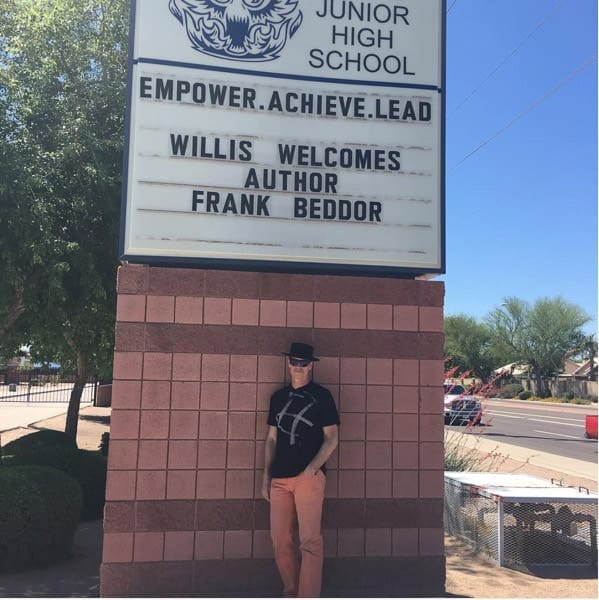 Beddor in a black hat and orange trousers standing beneath a
               junior high school sign that reads Willis Welcomes Author Frank
               Beddor.