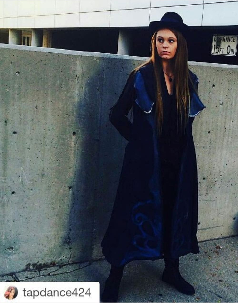 A young woman cosplaying Hatter Madigan in a black fedora and a
               long black coat lined and embroidered in blue,
               standing against a concrete wall.
