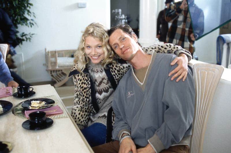 Beddor seated at a breakfast table on a set, leaning back
               while a young blonde woman in a leopard-print coat rests her chin
               on his shoulder, both smiling at the camera. A camera operator
               works in the doorway behind them.