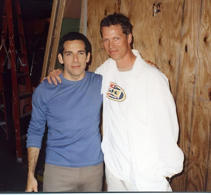Beddor backstage on a set, in a white sweatshirt and white
               trousers, an arm around a dark-haired man in a blue waffle shirt.
               Bare plywood flats, a stepladder and lighting stands are stacked
               behind them.