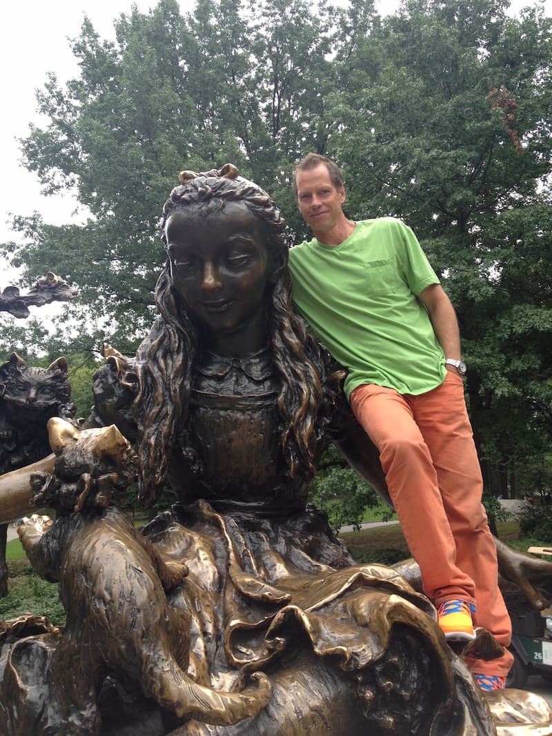 Beddor in a green T-shirt and orange trousers, climbed up onto
               the bronze Alice in Wonderland statue in Central Park with an arm
               around Alice&rsquo;s shoulders, the Cheshire Cat and the Dormouse
               beside them and summer trees behind.