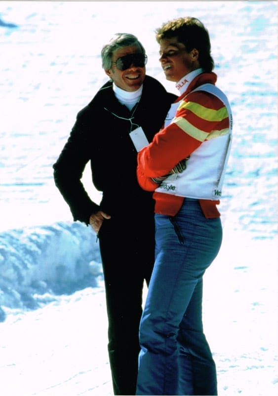 Beddor in a red and yellow race jacket on the snow, turned to
               his father, who stands beside him in a black ski suit and dark
               glasses with an event pass around his neck. Both are laughing.