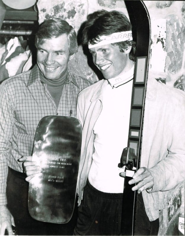 A black-and-white photograph of Beddor beside his father, both
               smiling. His father holds up an engraved plaque reading World Cup
               Freestyle, second place, men&rsquo;s ballet; Beddor, in a headband
               and an open jacket, holds his skis upright beside him.