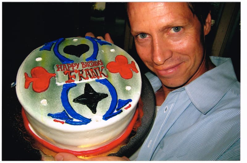 Beddor holding a birthday cake up beside his face, iced with
               the heart, club and diamond card suits of Wonderland around a
               banner reading Happy Birthday Frank.