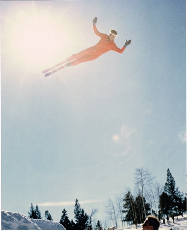 Beddor flat out in mid-air in a red one-piece suit, arms
               spread and skis together, high above the treeline with the sun
               flaring behind him.