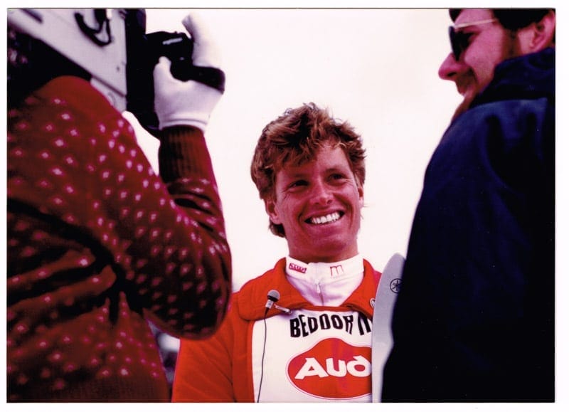 Beddor in the finish area being filmed for television, a
               camera operator in the foreground and a microphone at his chest.
               He is grinning; his bib reads Beddor III over an Audi logo.