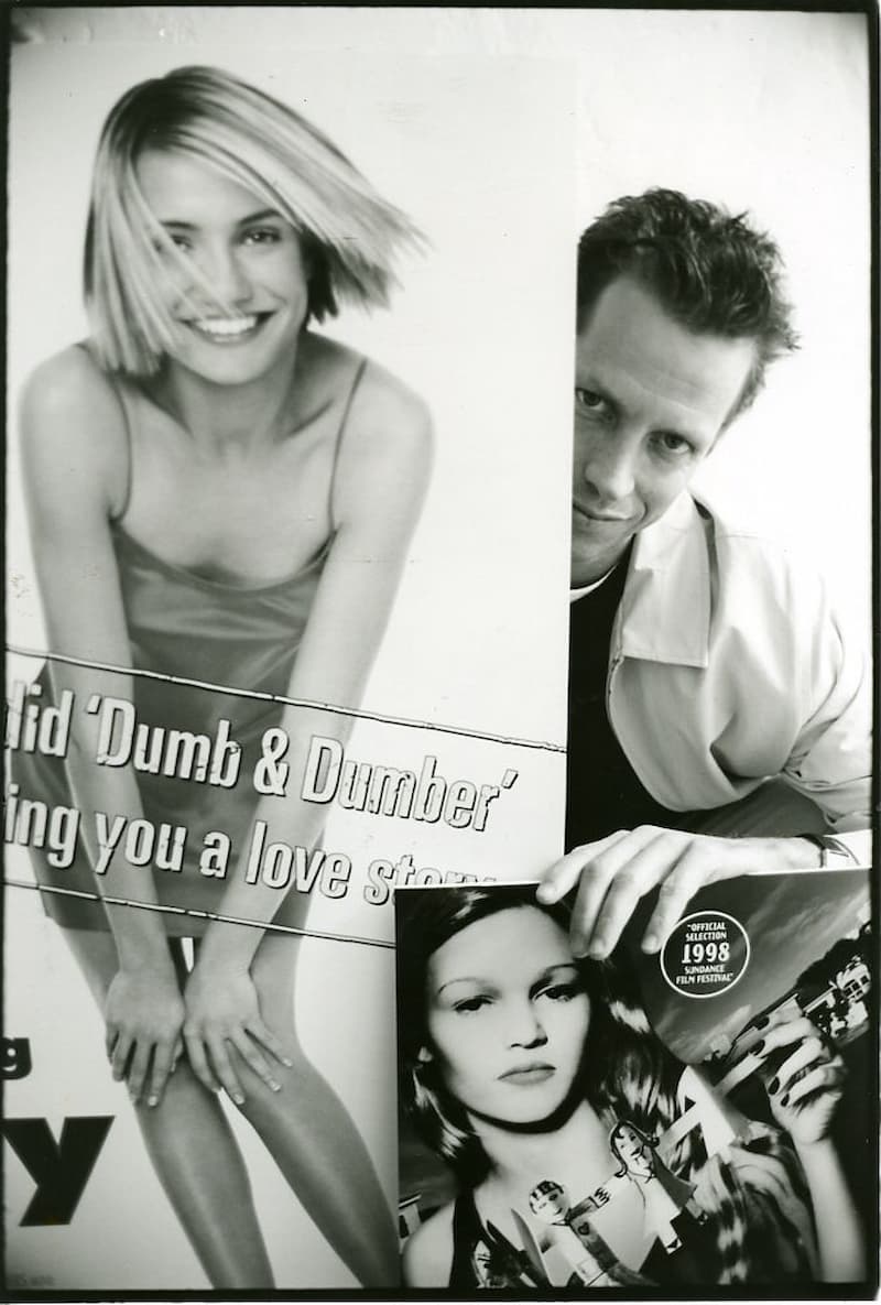 A black-and-white portrait from 1998: Beddor leaning in from
                    the right of the frame between two posters. Behind him is the
                    one-sheet for There&rsquo;s Something About Mary; in his
                    hand, held up to the camera, is the poster for Wicked with
                    its 1998 Sundance official selection badge.