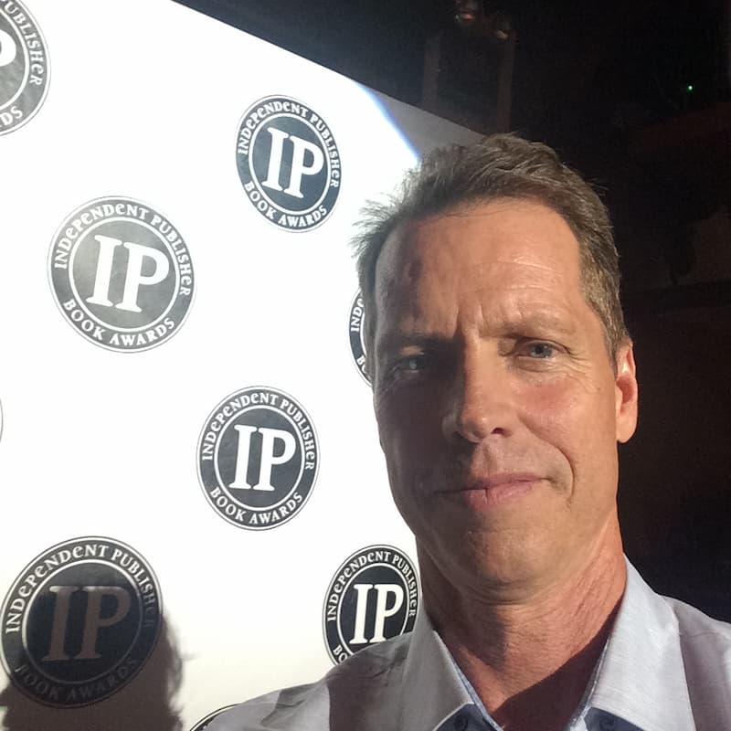 Beddor photographed at arm&rsquo;s length in front of a step
               and repeat banner printed with the Independent Publisher Book
               Awards seal.