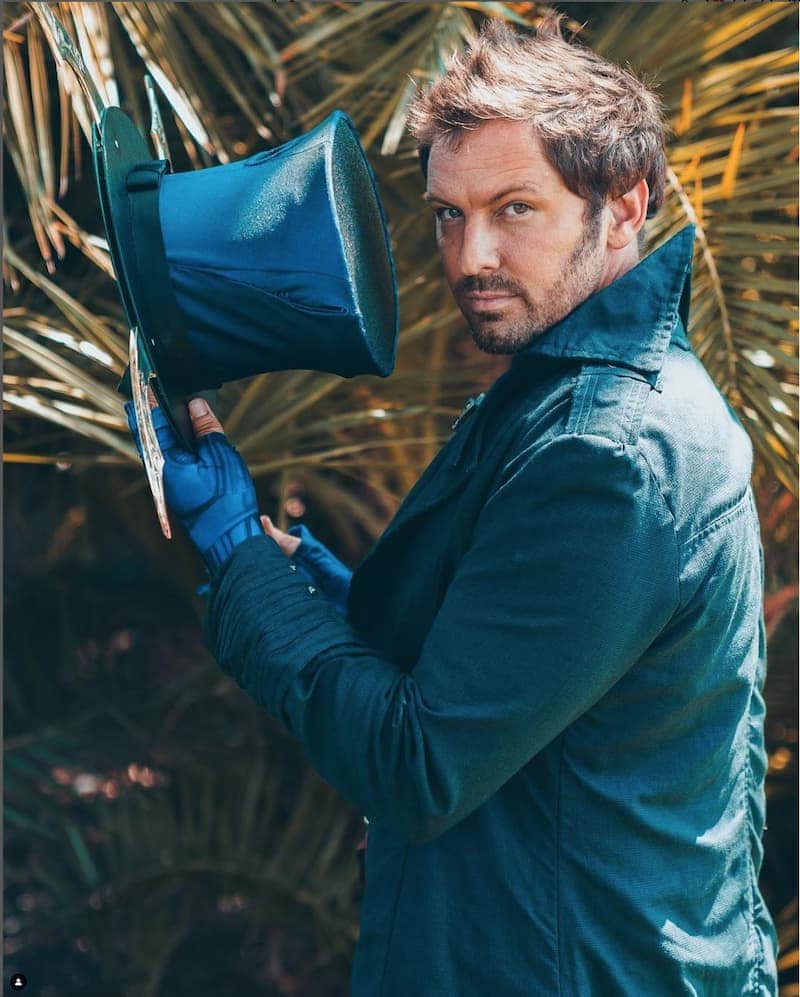 A Hatter Madigan cosplayer in a teal coat and blue gloves,
               glancing back over his shoulder and holding his
               top hat up beside him against palm fronds.