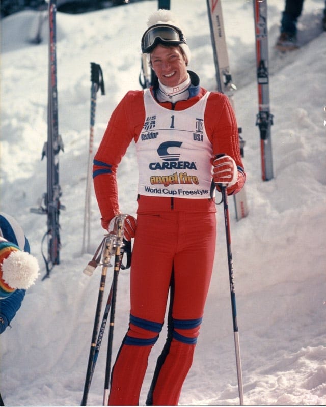 Frank Beddor in the start area in a red race suit, goggles
                    pushed up on his helmet and a pole in each hand, grinning.
                    The bib over his chest is number 1 and reads Beddor,
                    Carrera, Angel Fire, World Cup Freestyle.