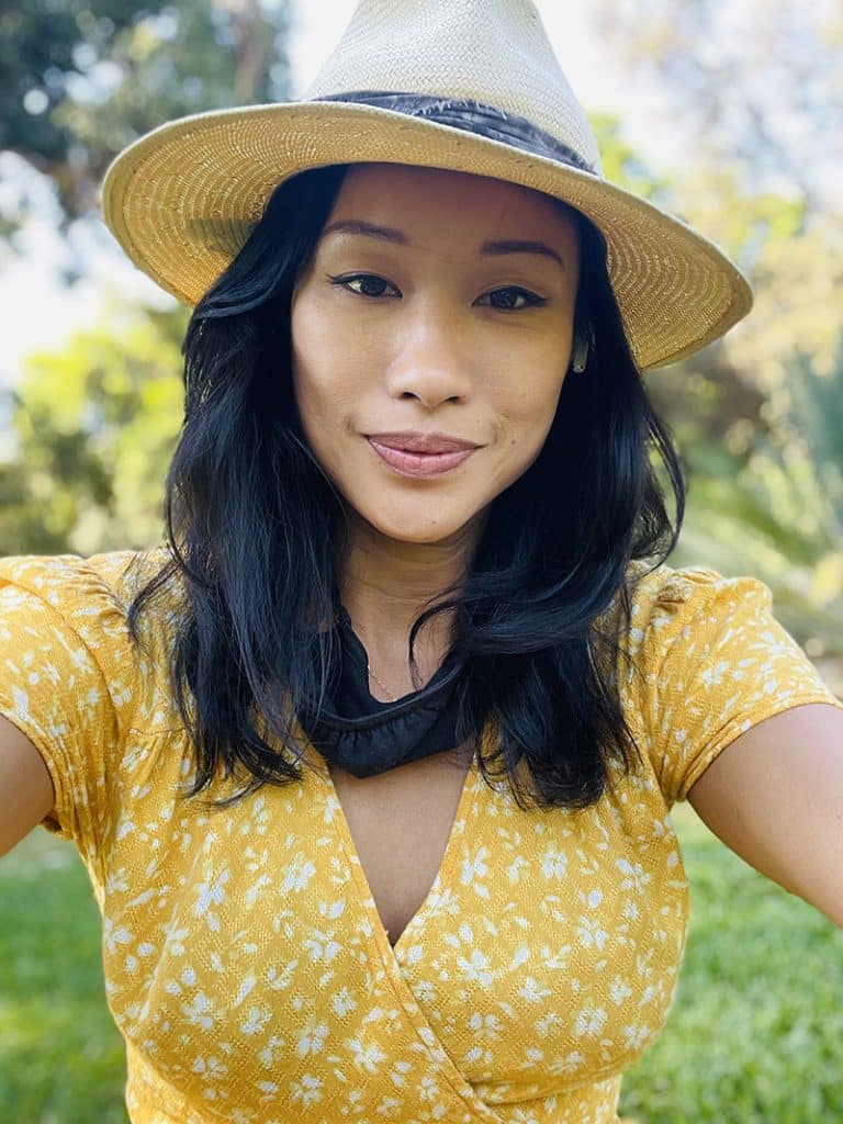 Teresa Lin outdoors in a straw hat and a yellow floral
                    dress, smiling at the camera, garden behind her.