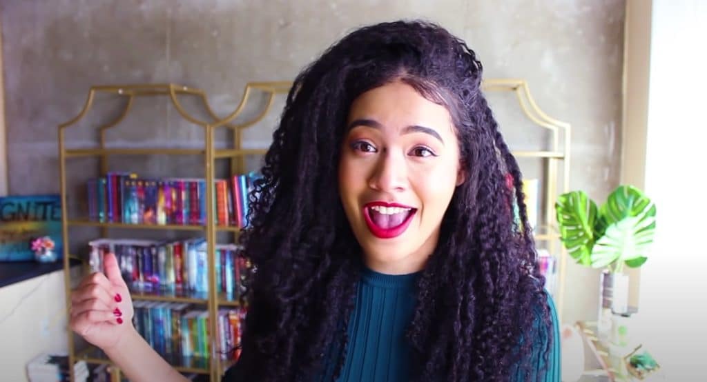 Booktuber NayaReadsandSmiles, with long, dark curly hair and red lipstick. She is wearing a green sweater and sitting in front of a large, gold bookshelf with a lot of books she has read. 
      