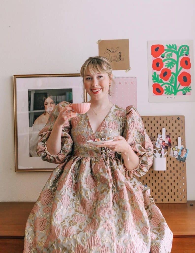 Booktuber: PeruseProject, in a dress holding a cup of tea and a small plate. She is standing in front of a wall with some art, a picture of green, red and black flowers, a painting of a woman and a bulletin board. 
      