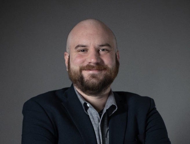 John Drain, bearded and smiling, in a dark jacket over a
                    grey shirt, against a plain grey backdrop.