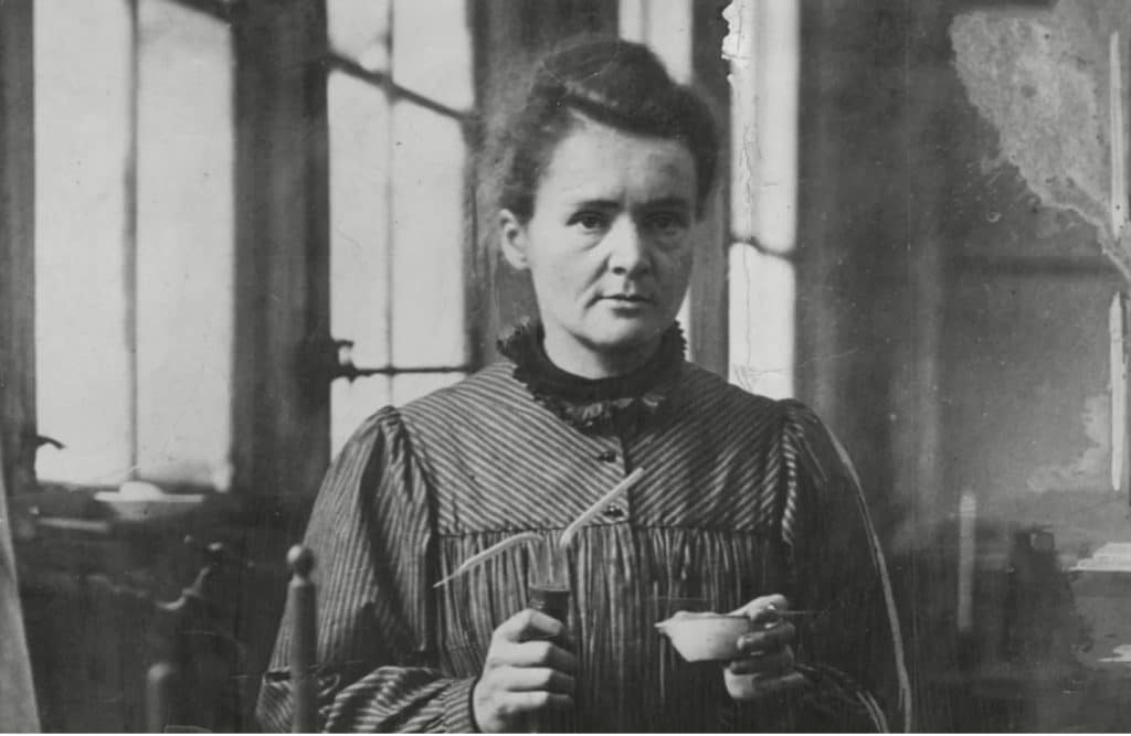 A historical photograph of Marie Curie in black and white. Her hair is in a bun as she holds scientific instruments 