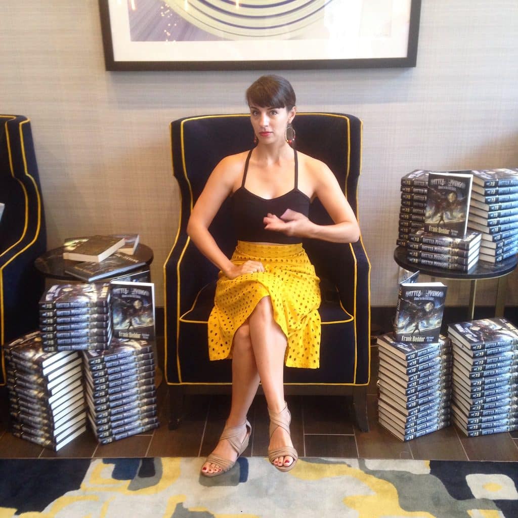 Author Adrienne Kress sitting down and surrounded by piles of the middle-grade novel "Hatter Madigan: Ghost in the H.A.T.B.O.X." by Frank Beddor and Adrienne Kress. 