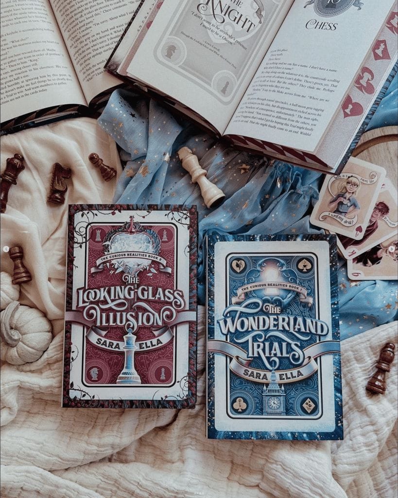 The Wonderland Trials" and "The Looking Glass Illusion" books by Sara Ella on a white and blue blanket surrounded by playing cards and chess pieces.