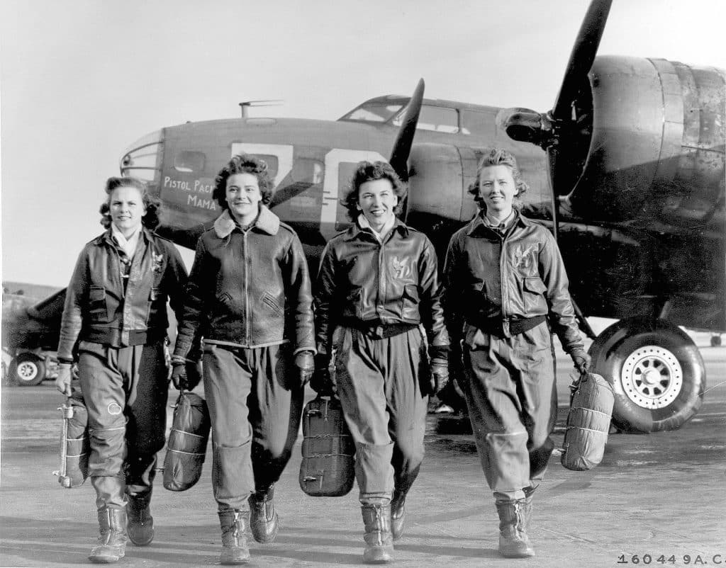 Four Women Airforce Service Pilots in flight jackets and
                      boots walking towards the camera with their parachutes,
                      a B-17 Flying Fortress behind them. United States Army
                      Air Forces photograph, 1944.