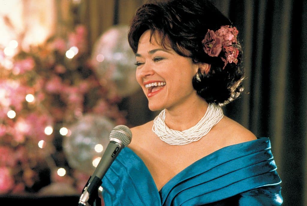Still image from the thriller movie "Wicked" featuring Linda Hart singing into a microphone in a blue dress and layered pearl necklace. 