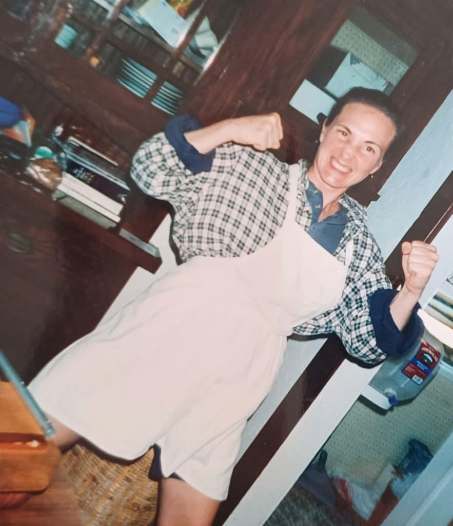 Liz Cavalier in a kitchen doorway in a white apron over a
                    checked shirt, both fists raised and grinning at the
                    camera.