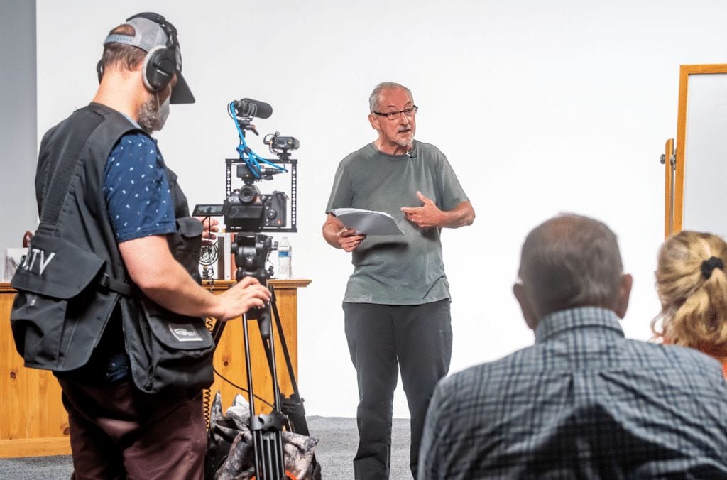 Photograph of actor and audiobook narrator Gerard Doyle speaking to a group of people in a classroom while being recorded by a camera. 