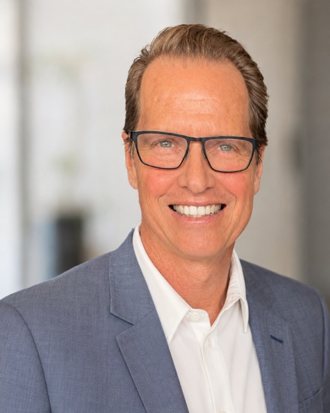 Frank Beddor in a blue-grey jacket over an open white shirt
                  and dark rectangular glasses, smiling straight at the camera.
                  The room behind him is thrown out of focus.