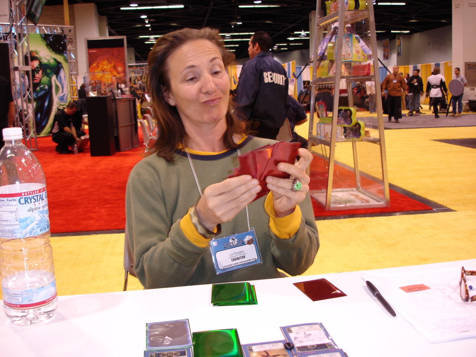 Liz Cavalier at an exhibitor table on a convention floor,
                    pulling a face as she folds a red foil card sleeve. Her
                    badge reads Liz Cavalier, Automatic Pictures, Exhibitor.