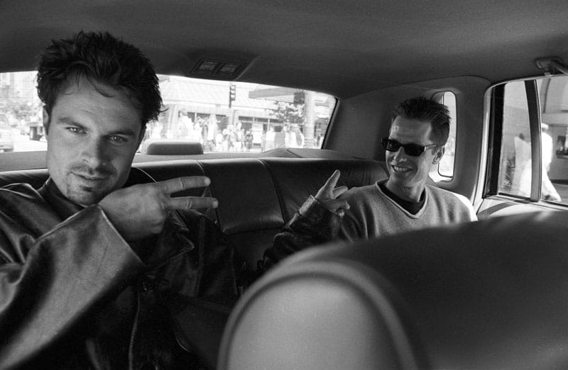 A black-and-white frame from the back seat of a car: Patrick
               Muldoon in a leather jacket on one side and Frank Beddor in
               sunglasses on the other, each pointing at the other across the
               bench, a street of shopfronts through the windows.