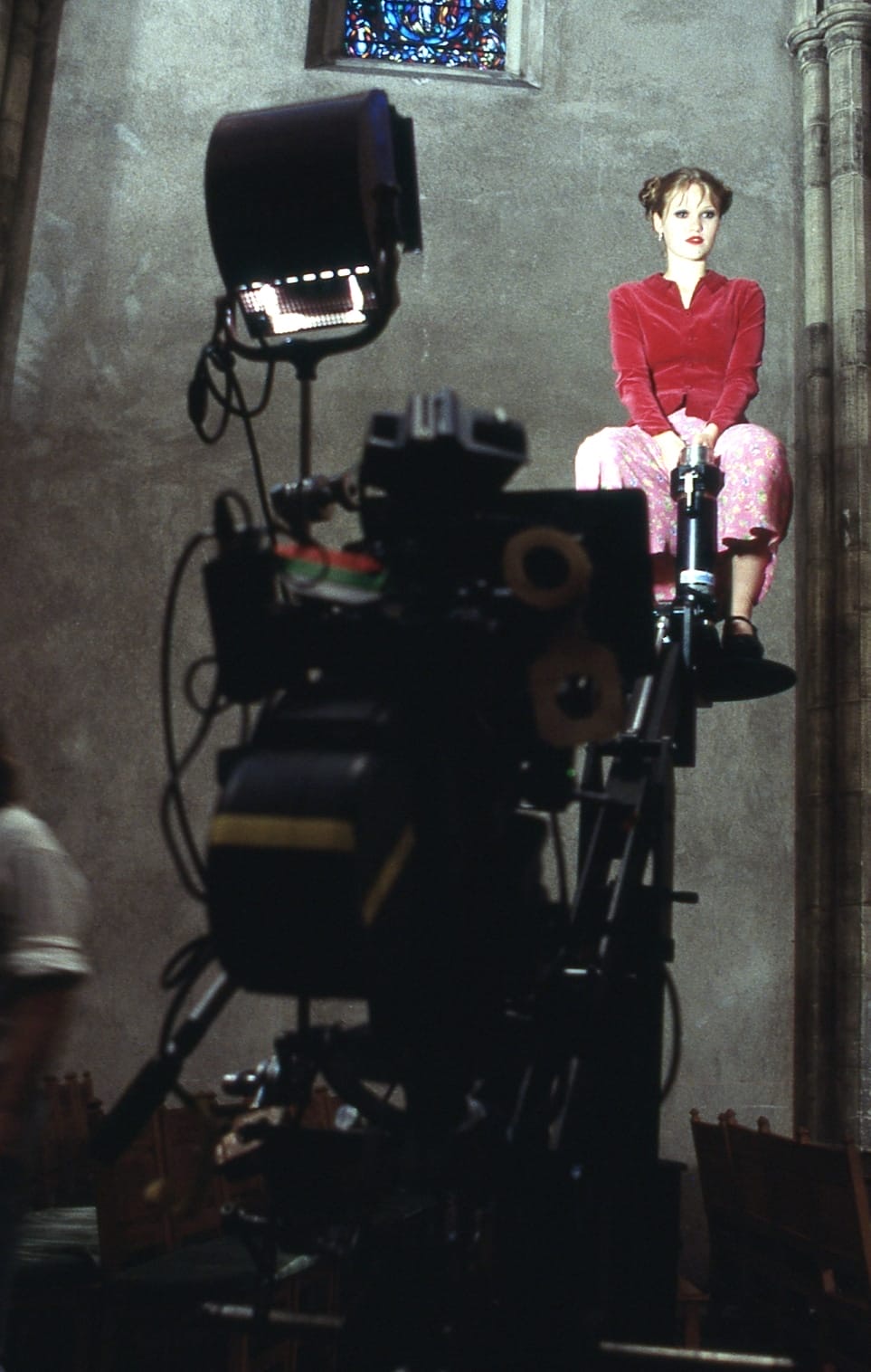 Julia Stiles perched high on the arm of a camera crane inside
               a church, in a red velvet jacket and pink floral trousers,
               waiting while the rig is set. A stained glass window and the
               lamp&rsquo;s open barn doors fill the wall behind her.