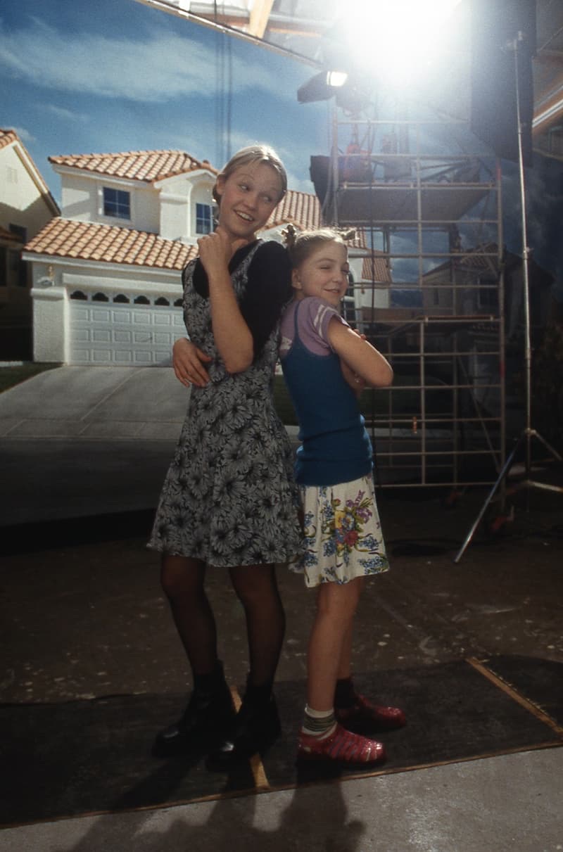 Julia Stiles and Vanessa Zima between takes, standing back to
               back and grinning over their shoulders, in front of the
               painted backdrop of a tile-roofed house that stands in for
               Casa Del Norte. Scaffolding and a lamp on a stand are just
               out of the shot behind them.